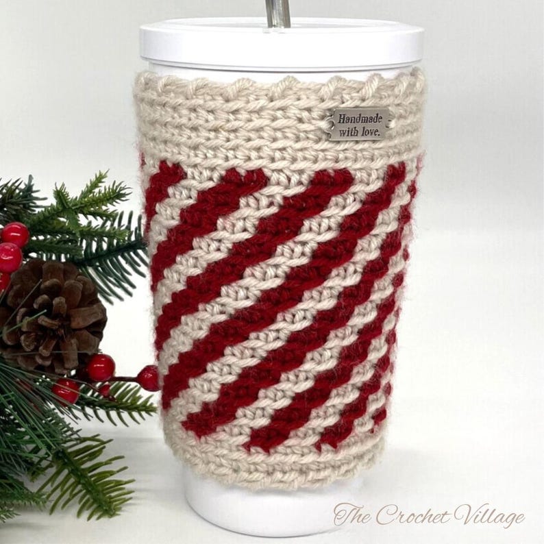 May include: A crocheted cup cozy in red and beige with a "Handmade with love." tag. The cozy is on a white travel mug.