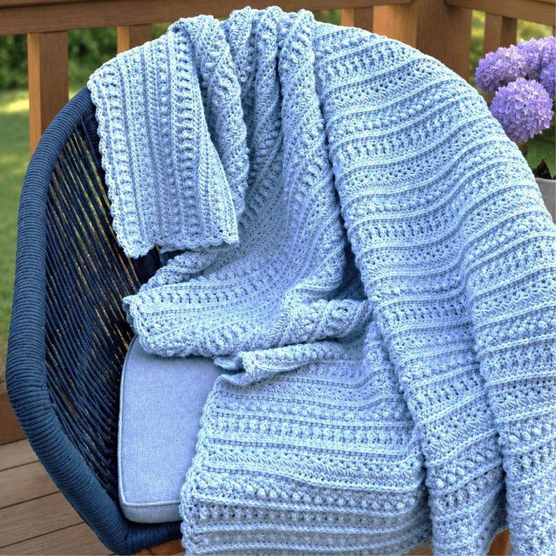 May include: A light blue crochet blanket featuring a textured pattern of alternating vertical stripes, including rows of raised bobble stitches and delicate shell-like motifs, draped over a dark blue woven chair with a light blue seat cushion.