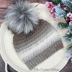 Puede incluir: Un gorro de punto de rayas marrones y blancas con un pompón de piel sintética gris. El gorro tiene una etiqueta de cuero marrón que dice "Handmade with Love".
