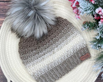 May include: A brown and white striped crocheted beanie with a gray faux fur pom pom. The beanie has a brown leather tag that says "Handmade with Love".