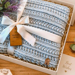 May include: A light blue crocheted blanket with a textured pattern of bobbles and ridges, wrapped in a white satin ribbon with a brown paper gift tag reading Handmade with love and a small wooden tag, presented in a white gift box with dried flowers.