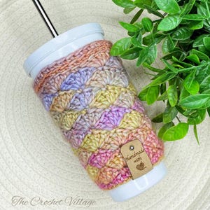 Puede incluir: Un vaso blanco con una pajita de acero inoxidable y una funda de ganchillo hecha a mano. La funda presenta un patrón de concha en tonos rosa, amarillo, melocotón y lavanda. Una pequeña etiqueta dice "Handmade with". El texto "The Crochet Village" está en la parte inferior.