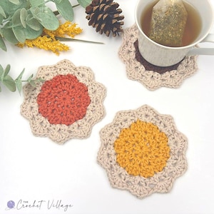 May include: Three round crochet coasters featuring a central circular motif in rust orange, mustard yellow, and dark brown, each surrounded by a scalloped beige border. One coaster supports a white mug containing tea with a visible tea bag.
