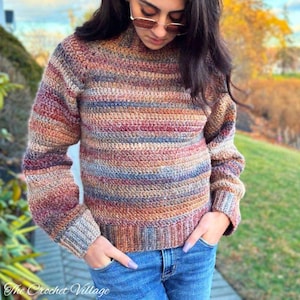May include: A handmade crochet sweater in a mix of warm colors, including brown, red, and blue stripes. The sweater has a ribbed hem and cuffs, and a high neckline. The text "The Crochet Village" is visible in the bottom left corner. The person is wearing blue jeans.