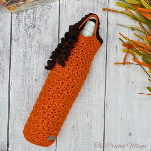 Puede incluir: Porta botellas de vino de crochet naranja con ribete de crochet marrón. El porta botellas está sobre un fondo de madera blanca.