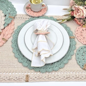 May include: A white cloth napkin with a scalloped edge and a light pink crocheted napkin ring, placed on a white plate. The plate sits on a rectangular beige crocheted placemat with a scalloped sage green border and a small brown leather tag.