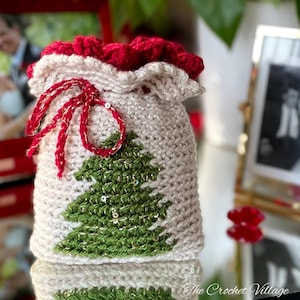 May include: A crocheted Christmas gift bag with a green Christmas tree design and gold sequins. The bag has a red drawstring and a red ruffled top.