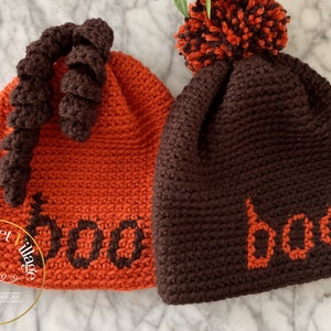 May include: Two handmade crochet Halloween beanies. One is orange with a brown curly accent and the word "boo" in brown. The other is brown with an orange pom-pom and the word "boo" in orange. The Crochet Village logo is in the bottom left.