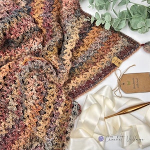 May include: A crocheted blanket featuring a variegated yarn pattern in shades of rust, brown, cream, and charcoal. A small rectangular tag reads Handmade with love. A gold-toned crochet hook and a white satin ribbon are placed near the blanket.