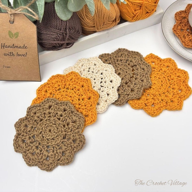 May include: A set of five round, scalloped-edge crochet coasters in shades of brown, tan, and orange. A brown paper tag with the text "Handmade with love!" is attached. The coasters are arranged in a curved line on a white surface.