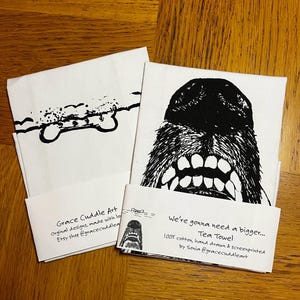 May include: Two tea towels with black and white illustrations. The first towel features a dog bone with clouds above it. The second towel features a dog's face with the text "We're gonna need a bigger... Tea Towel 100% cotton, hand drawn & screenprinted by Sonia @gracecuddleart".