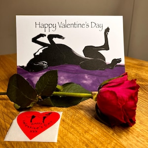 May include: A Valentine's Day card featuring a black and white illustration of a dog lying on a purple blanket. The card reads "Happy Valentine's Day". A red rose and a red heart sticker with the text "Happy Valentine's Day" are also in the image.