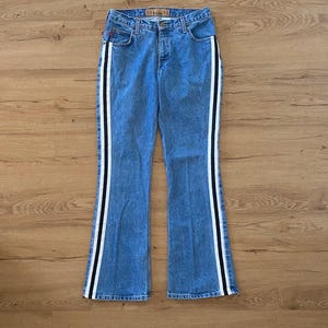 May include: Light blue denim jeans with a flared leg and a white and black stripe down the side. The jeans have a classic five-pocket design and a button and zipper closure. The jeans are made of a medium-wash denim.