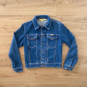 May include: A blue denim jacket with a button-up front, two chest pockets, and two side pockets. The jacket has a classic collar and the brand name "RUSTLER" is visible on the label. The jacket is made of a sturdy denim material.