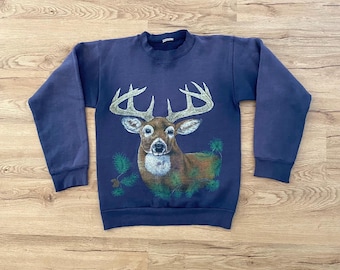 May include: A navy blue sweatshirt featuring a detailed graphic of a deer with large antlers. The deer is brown and white, set against a backdrop of green foliage. The sweatshirt has long sleeves and a crew neck.