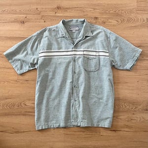 May include: A short-sleeved button-down shirt in a light blue and white horizontal pinstripe pattern. The shirt features a camp collar, a single chest pocket on the left side, and a horizontal white stripe across the chest with the Rusty brand label.