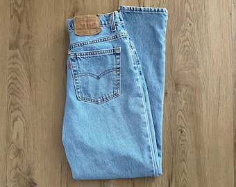 May include: Light blue denim jeans with a classic five-pocket design. The jeans feature a brown leather patch on the back and a red tag on the back pocket. The jeans are folded neatly on a wooden surface.