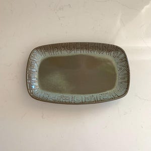 May include: Rectangular ceramic serving tray with a raised border featuring an abstract design. The tray has a muted green center and a brown and blue-green border. The tray is likely used for serving food or displaying decorative items.