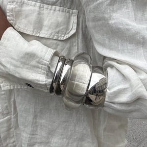 Könnte beinhalten: Ein cremefarbenes Leinenhemd mit zwei Vordertaschen und langen Ärmeln. Das Hemd wird mit einem Satz silberner Armreifen getragen. Die Armreifen sind unterschiedlich breit und haben eine reflektierende Oberfläche. Das Hemd ist oben aufgeknöpft.