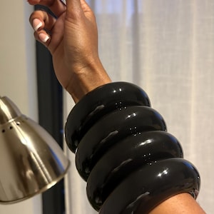 Peut inclure: Une pile de cinq bracelets noirs brillants sur un poignet. Les bracelets épais et arrondis ont une finition très brillante. Le bras est tendu et la main est ouverte avec une manucure française.