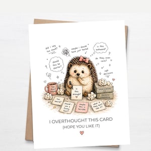 May include: A greeting card featuring a cartoon hedgehog with a pink bow, surrounded by thought bubbles and notes. The card reads "I OVERTHOUGHT THIS CARD (HOPE YOU LIKE IT)". The illustration includes a coffee mug and small books.