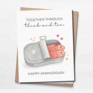 Peut inclure: Carte de vœux avec une illustration à l'aquarelle de deux poissons dans une boîte de sardines. La carte porte l'inscription "Together through thick and tin" et "Happy Anniversary". La carte est sur une enveloppe brune.
