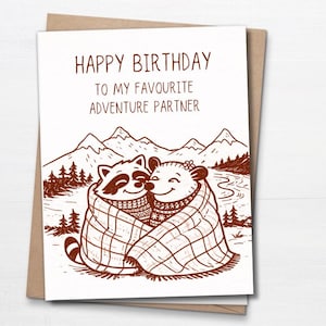 Peut inclure: Carte d'anniversaire avec une illustration marron d'un raton laveur et d'un ours sous une couverture. Le texte dit : "JOYEUX ANNIVERSAIRE À MON PARTENAIRE D'AVENTURE PRÉFÉRÉ" avec un arrière-plan de montagne et de forêt.