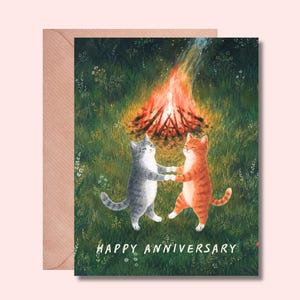 Op de afbeelding: Een wenskaart met een illustratie van een grijze cyperse kat en een oranje cyperse kat die elkaars pootjes vasthouden in een grasveld voor een fel kampvuur. De tekst "HAPPY ANNIVERSARY" staat onderaan in witte letters gedrukt.