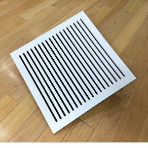 May include: White square vent cover with a grid of vertical slots. The vent cover is lying on a wood floor.