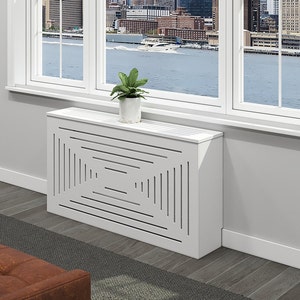 May include: White radiator cover with a geometric design. The cover has a rectangular shape and is made of wood. It is sitting on a wooden floor in front of a window.
