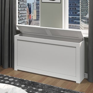 May include: White radiator cover with a slatted lid, open to reveal the radiator. The cover is in a living room setting with a window and curtains in the background.