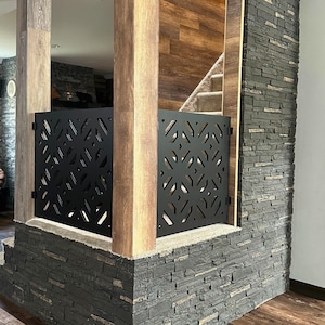 May include: Black metal decorative gate with a geometric pattern, installed between wooden posts and a stone wall. The gate is a safety feature, likely for a staircase or doorway, and adds a modern touch to the interior.