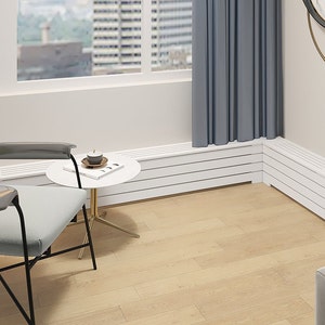May include: A white radiator cover with a slatted design is installed beneath a window in a modern living room. The radiator cover is in the corner of the room and extends along two walls. A gray chair with black metal legs sits in front of the radiator cover. A small round table with a white top and gold legs sits next to the chair. The table has a book and a candle on it.