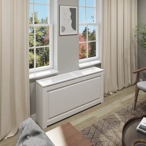 May include: White radiator cover with a slatted top and a decorative trim. The cover is in a living room setting with a rug and a coffee table.