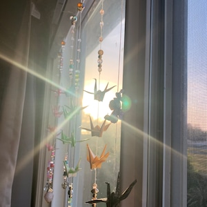 Könnte beinhalten: Ein Fenster mit Sonnenlicht, das durchscheint. An dem Fenster hängen Fäden mit Perlen und Origami-Papierkranichen in verschiedenen Farben, darunter Grün, Blau, Gold und Schwarz.