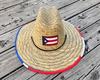 May include: A straw hat with a red, white, and blue trim and a Puerto Rican flag patch. The hat is sitting on a wooden surface.