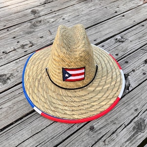 Puede incluir: Un sombrero de paja con un borde rojo, blanco y azul y un parche de la bandera puertorriqueña. El sombrero está sobre una superficie de madera.
