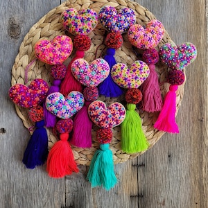 May include: A collection of ten heart-shaped ornaments, each featuring vibrant, multi-colored floral embroidery. Every heart is attached to a small, textured pom-pom and a long, flowing tassel in bright colors including purple, pink, red, green, and teal.