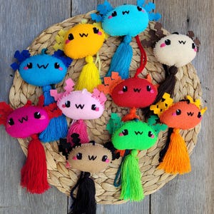 May include: A collection of ten felt axolotl ornaments in various colors including blue, yellow, pink, red, green, orange, and brown. Each plush figure features embroidered facial details, a w-shaped mouth, and a matching hanging tassel.