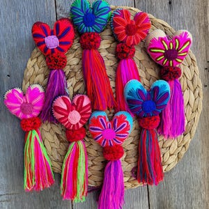 May include: Eight heart-shaped ornaments featuring vibrant embroidered floral patterns in shades of pink, red, blue, and green. Each heart is attached to a small red pom-pom and a long, multi-colored yarn tassel in bright neon and primary colors.