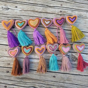 May include: A collection of 14 colorful heart-shaped ornaments with tassels. Each heart is made of yarn and has a different color combination. The tassels are also made of yarn and come in a variety of colors.