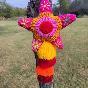 May include: A star-shaped textile ornament in vibrant fuchsia, featuring intricate multi-colored floral embroidery and a central circular motif. A cascading series of four layered tassels in yellow, red, orange, and dark red hangs from the bottom.