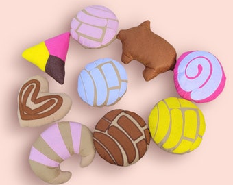 May include: A set of nine colorful felt pillows shaped like various Mexican pastries. The pillows are in shades of brown, pink, yellow, blue, and white.