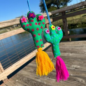 May include: Two green felt cactus ornaments with pink and yellow flowers and tassels. The cactus on the left has a yellow tassel and the cactus on the right has a pink tassel.
