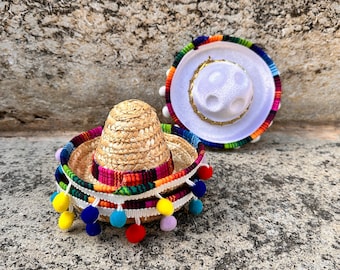 May include: Two miniature sombreros, one straw and one white with colorful trim, with colorful pom-poms around the base.