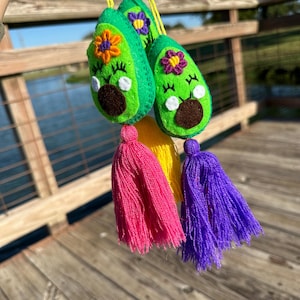 May include: Three green felt avocado ornaments with embroidered details, including a flower and a face. Each avocado has a colorful tassel hanging from the bottom.