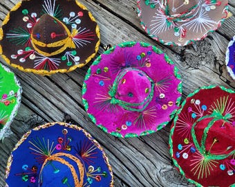 May include: A collection of miniature sombreros in various colors including orange, brown, pink, blue, red, and black. Each hat features intricate embroidery with radiating thread patterns, colorful sequins, and a braided trim along the brim edge.