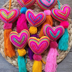 May include: A collection of seven heart-shaped ornaments made of multi-colored yarn. Each heart features concentric stripes in vibrant shades of pink, orange, yellow, blue, and green. Each ornament includes a matching yarn tassel and a pom-pom.
