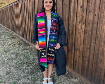 May include: A person wearing a black graduation gown and holding two colorful stoles. One stole is striped with the text "Class of 2026", and the other has the text "Educated & Unstoppable". The person is wearing white sandals.