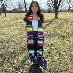 May include: A woman wearing a colorful serape scarf with a black background and stripes of red, orange, yellow, green, and blue. The scarf is fringed at the ends. She is wearing a white top and floral print pants.